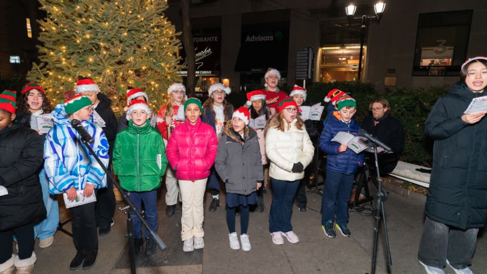A children's choir carol singing beside the garden's lit Christmas tree